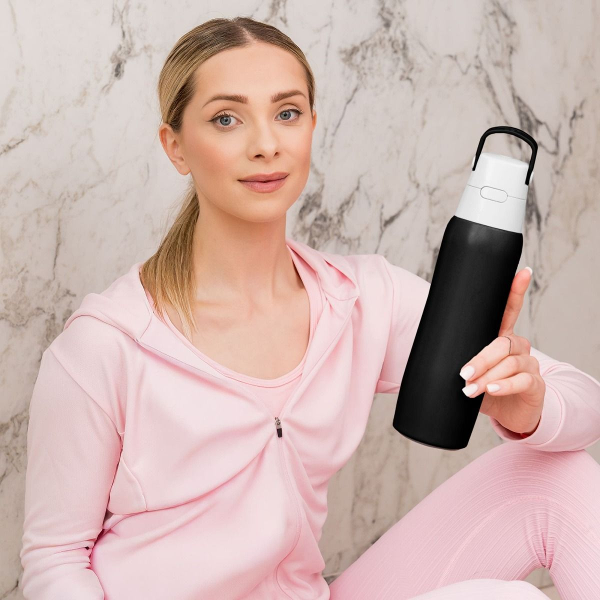 Blondynka w różowym stroju sportowym trzyma czarno-białą butelkę z wodą. Siedzi przed marmurową ścianą.