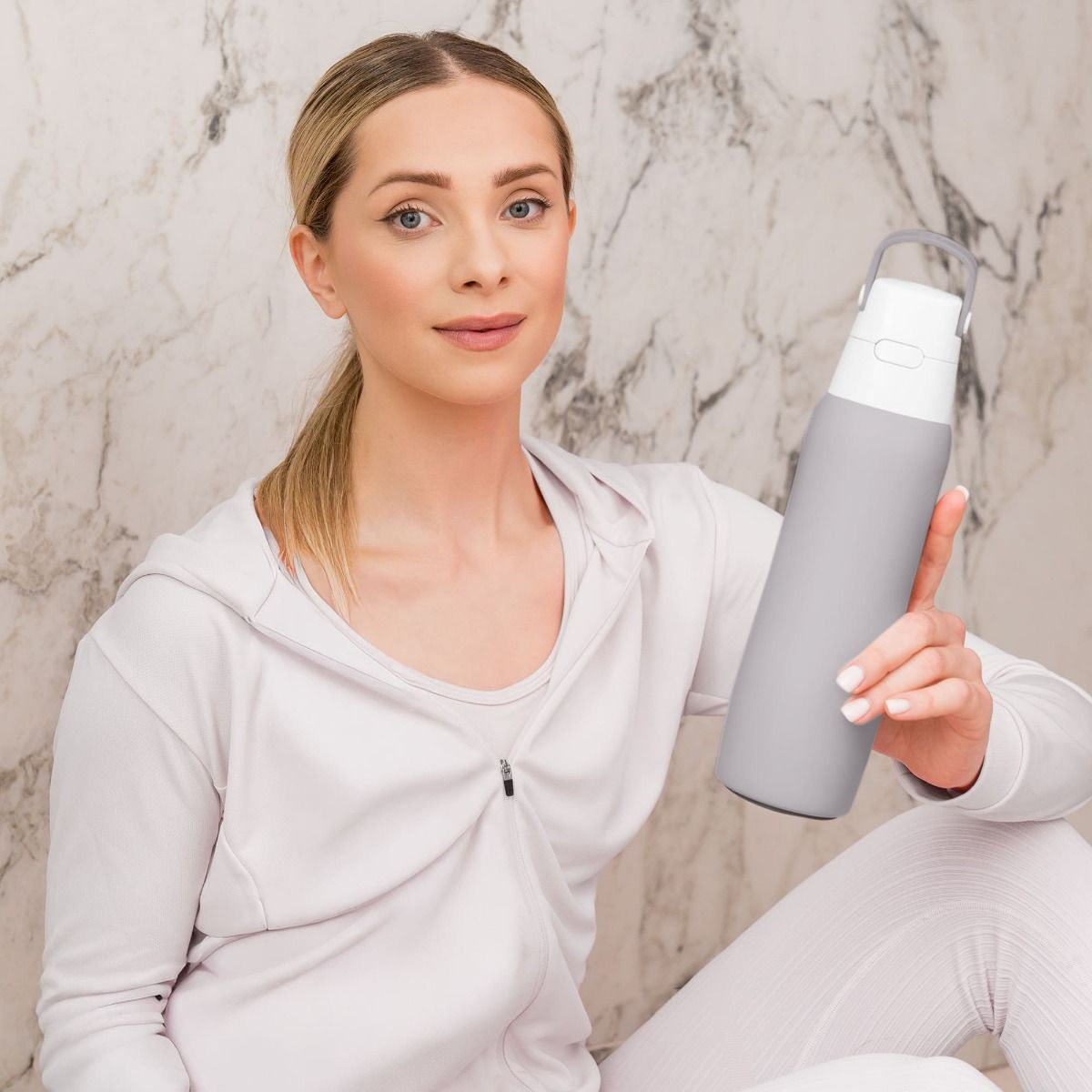 Kobieta w białym stroju sportowym trzyma szarą butelkę z wodą. Ma blond włosy związane w kucyk i uśmiecha się.