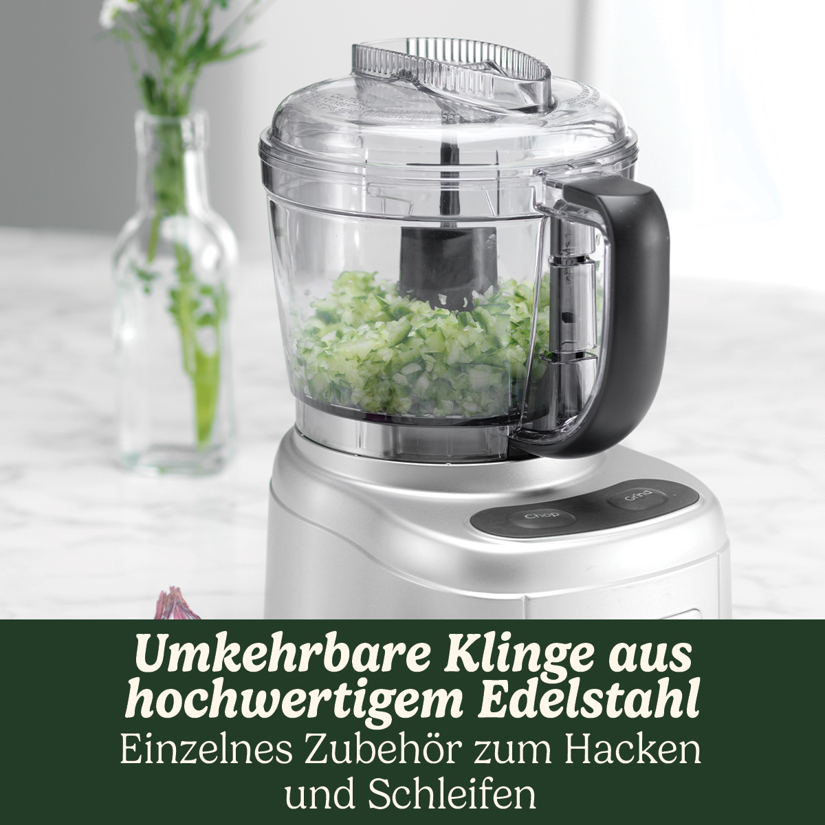 Ein silberner Foodprocessor mit gehacktem, grünem Gemüse. Im Hintergrund eine Vase mit Blumen.