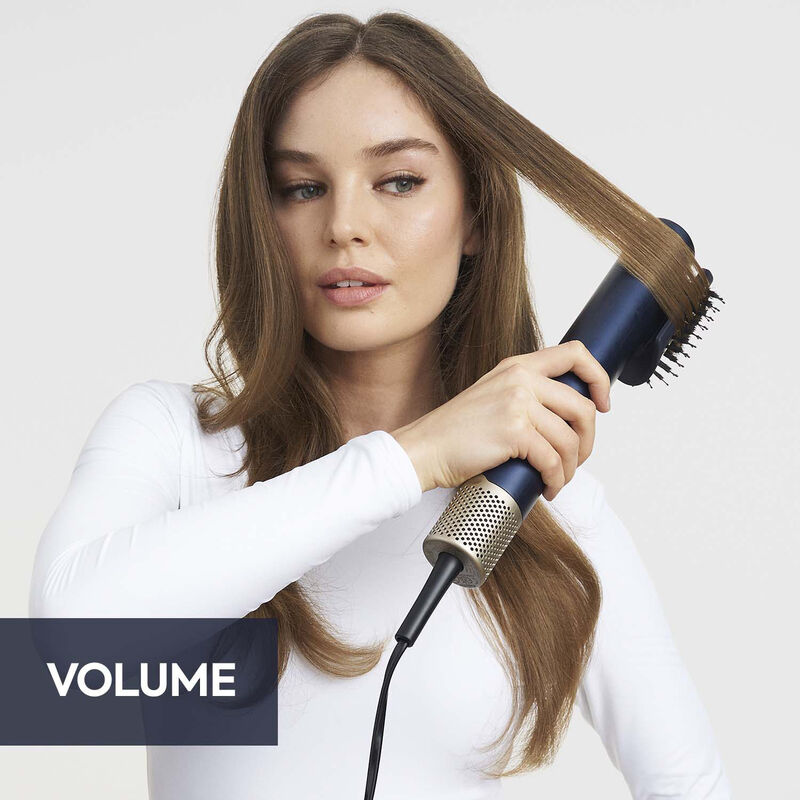 Eine Frau in einem weißen Shirt benutzt einen blau-goldenen Haarstyler für ihr braunes Haar. Der Hintergrund ist weiß, und das Wort VOLUME ist zu sehen.