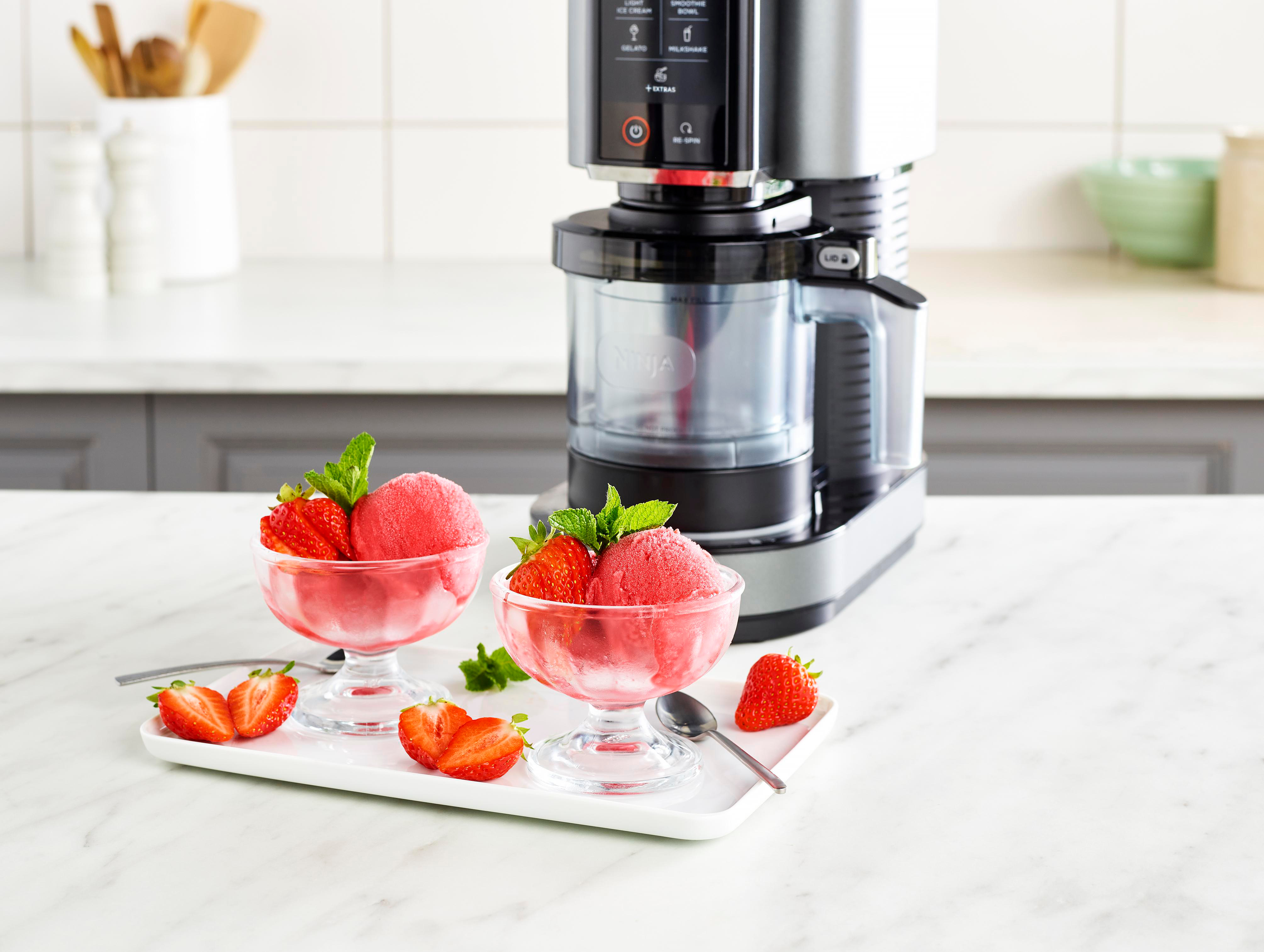 Erdbeersorbet in Glasschalen mit Minze. Im Hintergrund ein Mixer. Auf einem Tablett geschnittene Erdbeeren.