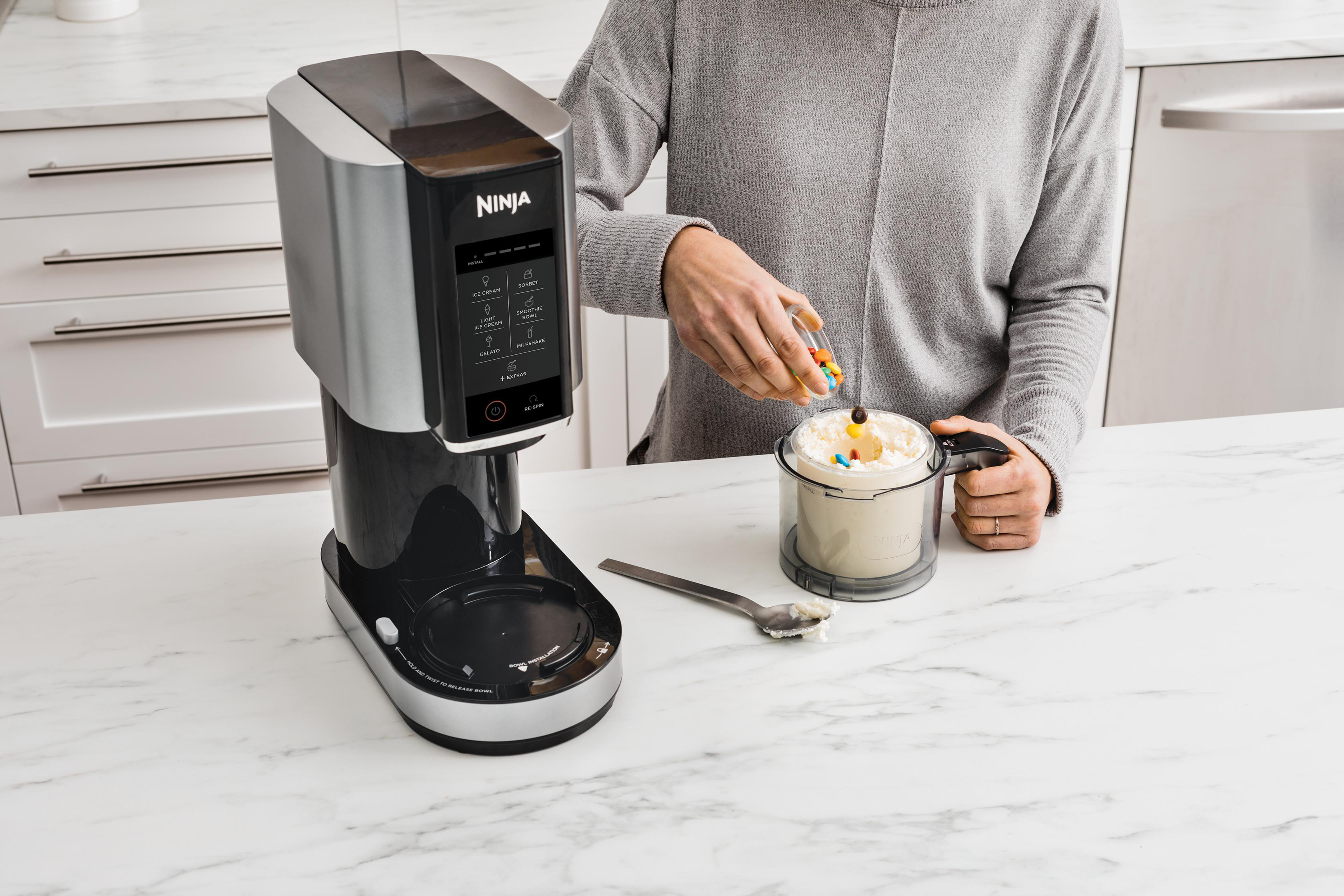 Eine Person fügt Süßigkeiten zu einem Dessert in einer Ninja-Maschine hinzu. Ein Löffel mit Eiscreme liegt auf der Theke. Der Hintergrund zeigt eine Küche.