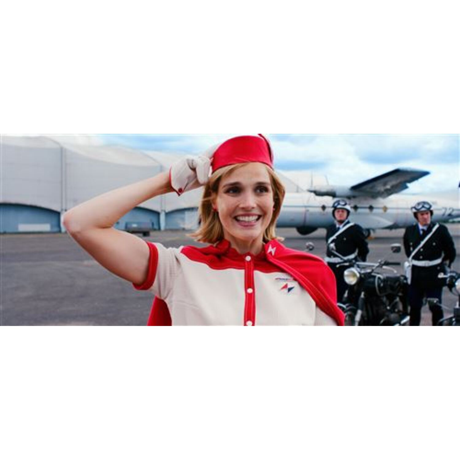 Een vrouw in een rood en wit uniform groet. Achter haar staan twee mannen in uniform op motorfietsen en een vliegtuig.