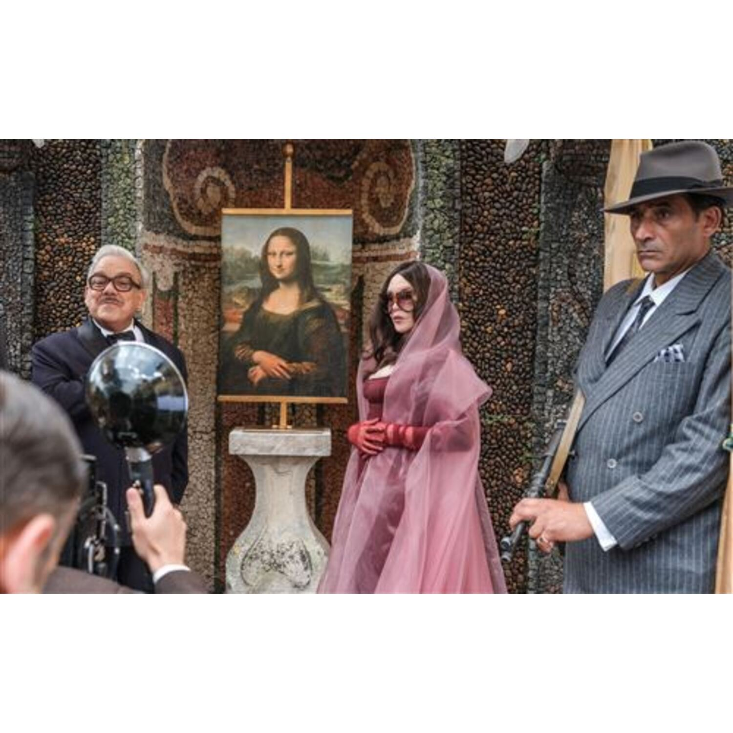 Mensen staan bij een schilderij van de Mona Lisa. Een vrouw in het roze, een man in een grijs pak en een man met een camera zijn zichtbaar.