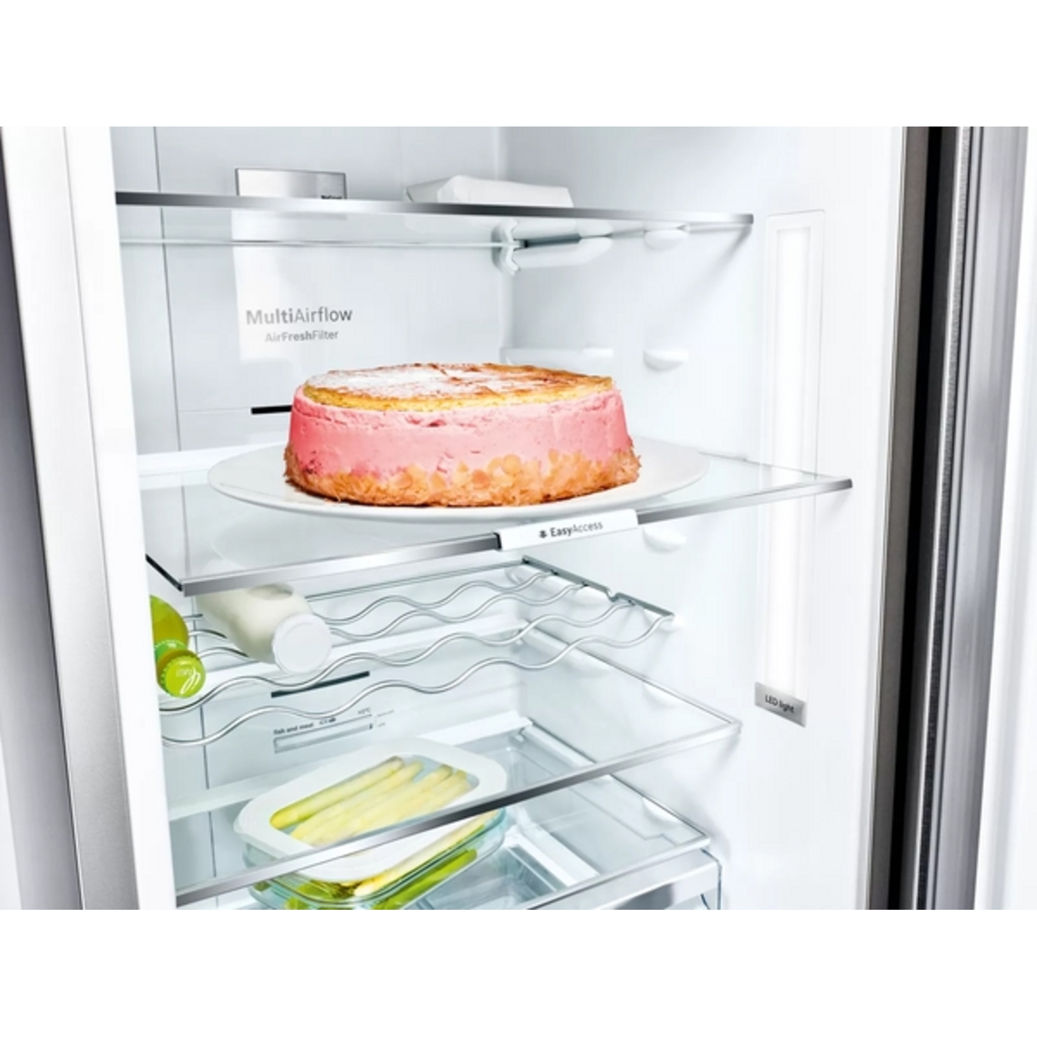 In een koelkast staat een roze cake op een bord. Andere items zijn flessen, asperges en een lamp.