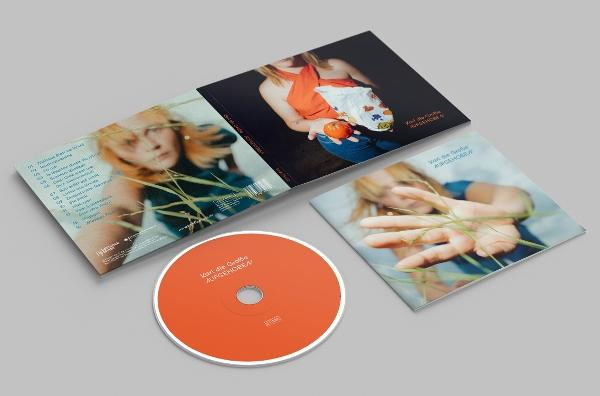CD-Album mit einer Frau auf dem Cover. Das offene Album, die CD und die Hülle liegen auf einer grauen Oberfläche. Das Album hat orange und weiße Farben.