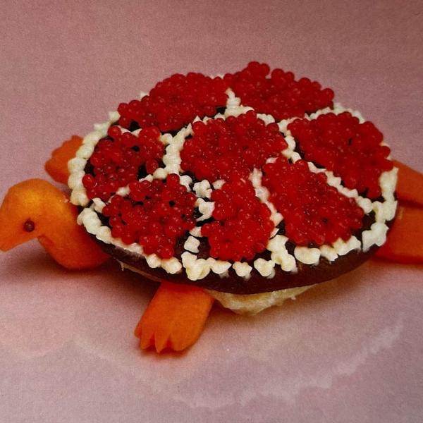 Eine Schildkröte aus Essen. Roter Kaviar oben, weiße Sahne darum. Orangefarbene Karottenbeine und -kopf. Rosa Hintergrund.