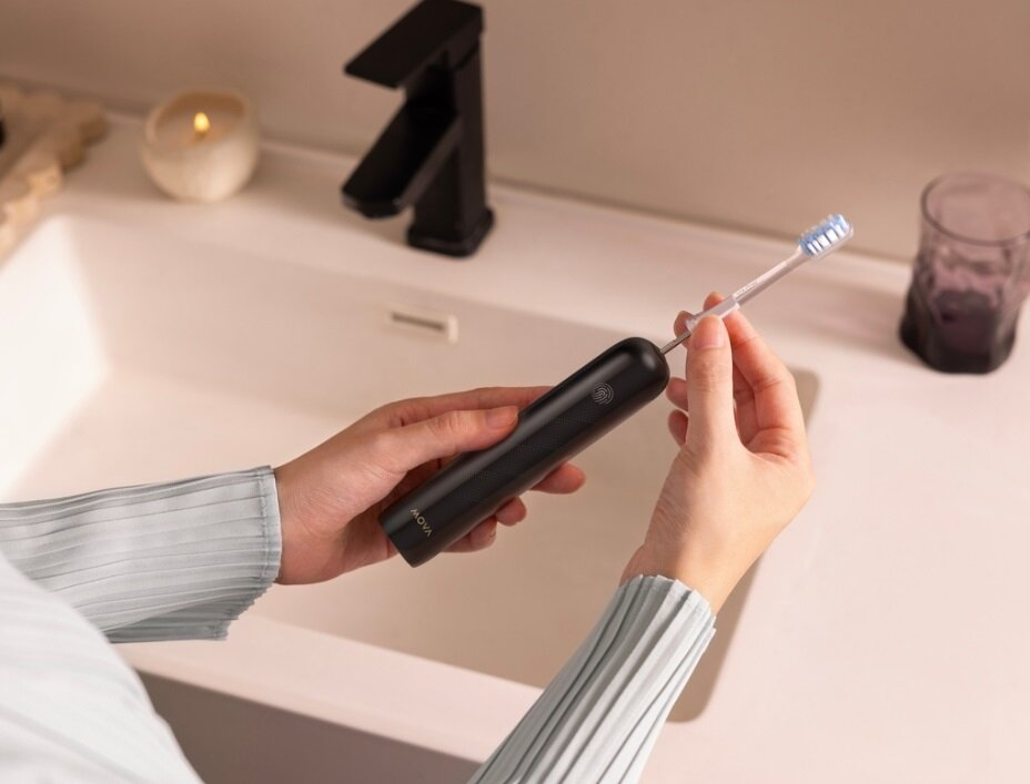 Ręce trzymające czarną elektryczną szczoteczkę do zębów i główkę szczoteczki. Umywalka, kran, świeca i szklanka w tle.