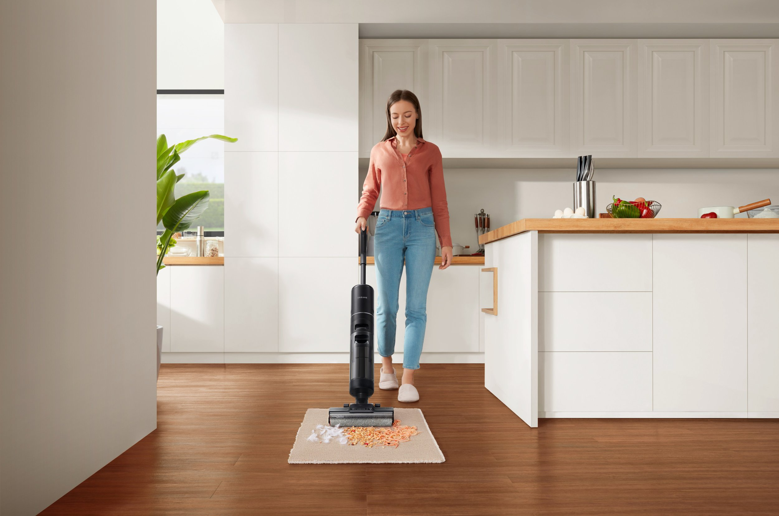 Kobieta używa czarnego odkurzacza w kuchni. Odkurzacz jest nad dywanem ze śmieciami. Białe szafki w tle.