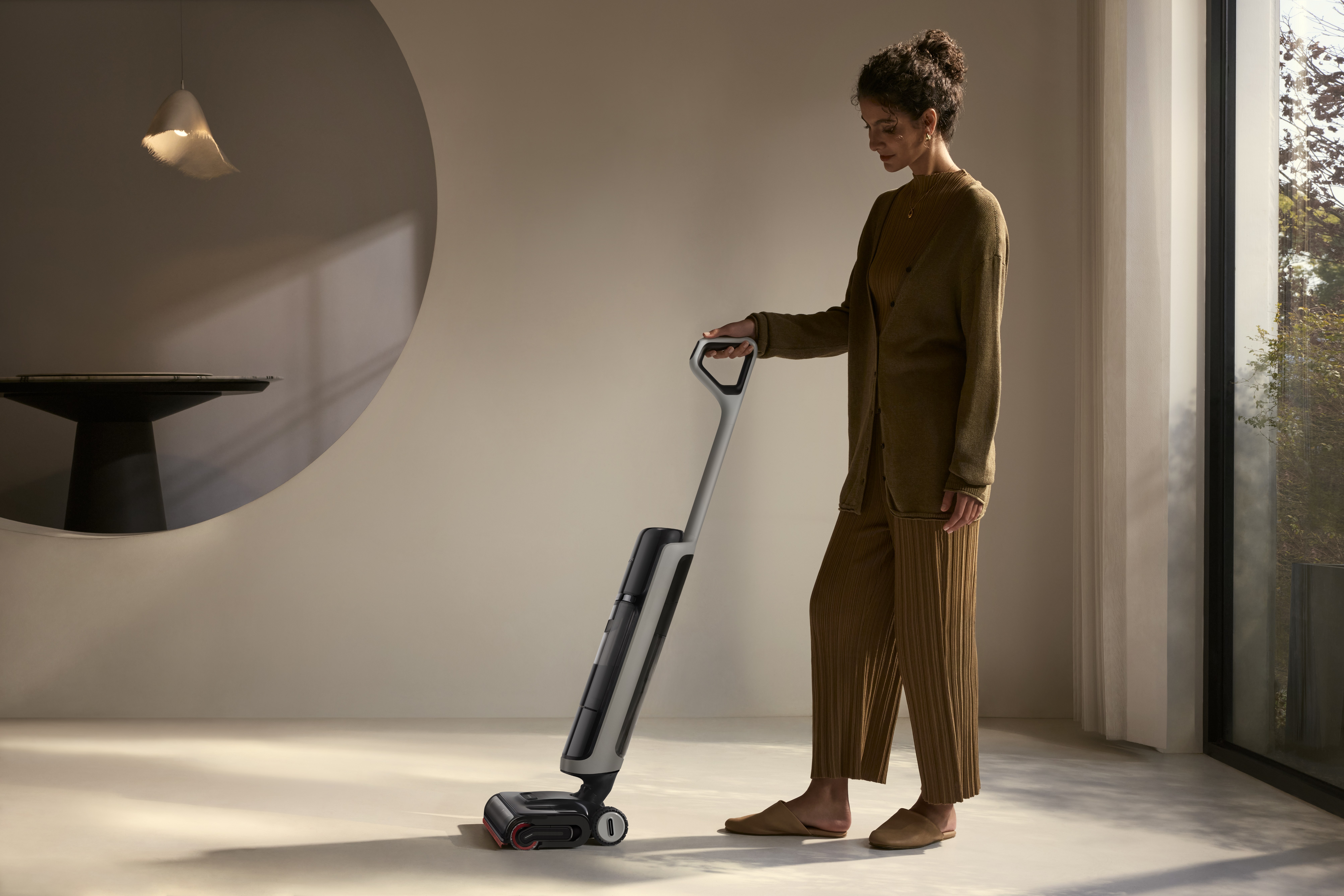 Kobieta używa odkurzacza. Nosi brązowe ubrania. Duże okno i okrągły wzór są w tle.