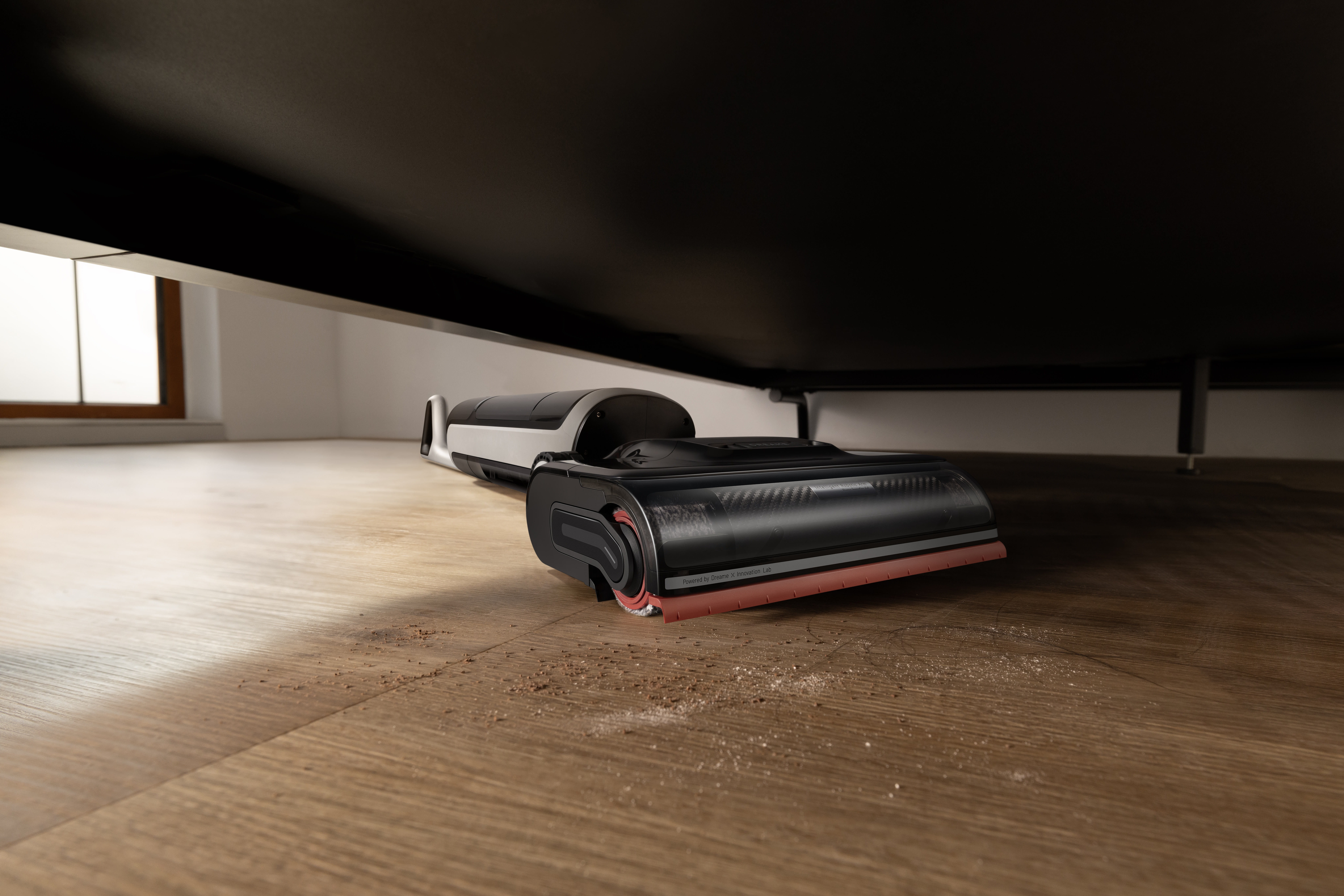 Czarno-biały odkurzacz stoi na drewnianej podłodze. Odkurzacz jest pod czarnym stołem. Na podłodze leżą zanieczyszczenia.