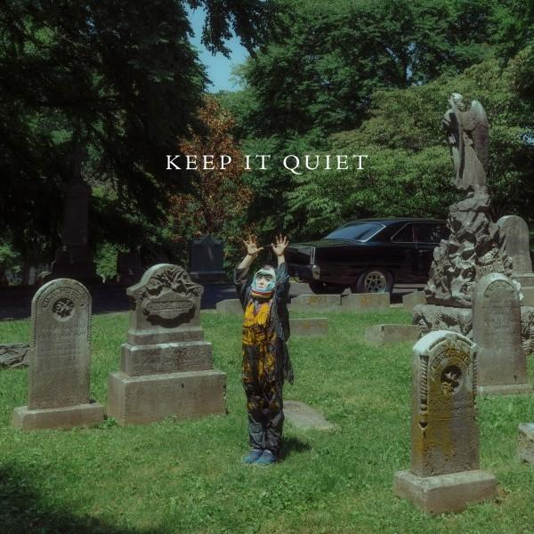Person im Kostüm hebt die Hände auf einem Friedhof, Text 'KEEP IT QUIET', schwarzes Auto, Grabsteine, Bäume.