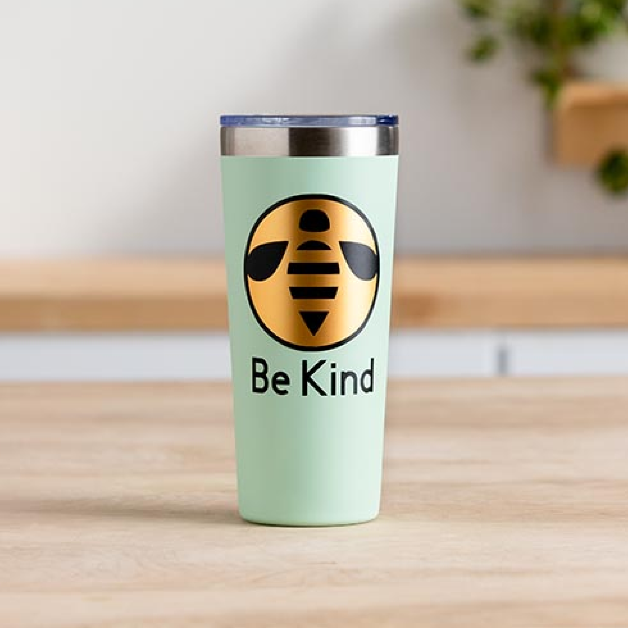 Ein hellgrüner Becher mit einem Bienenlogo und dem Text 'Be Kind'. Auf einer Holzoberfläche mit verschwommenem Hintergrund einer Pflanze platziert.
