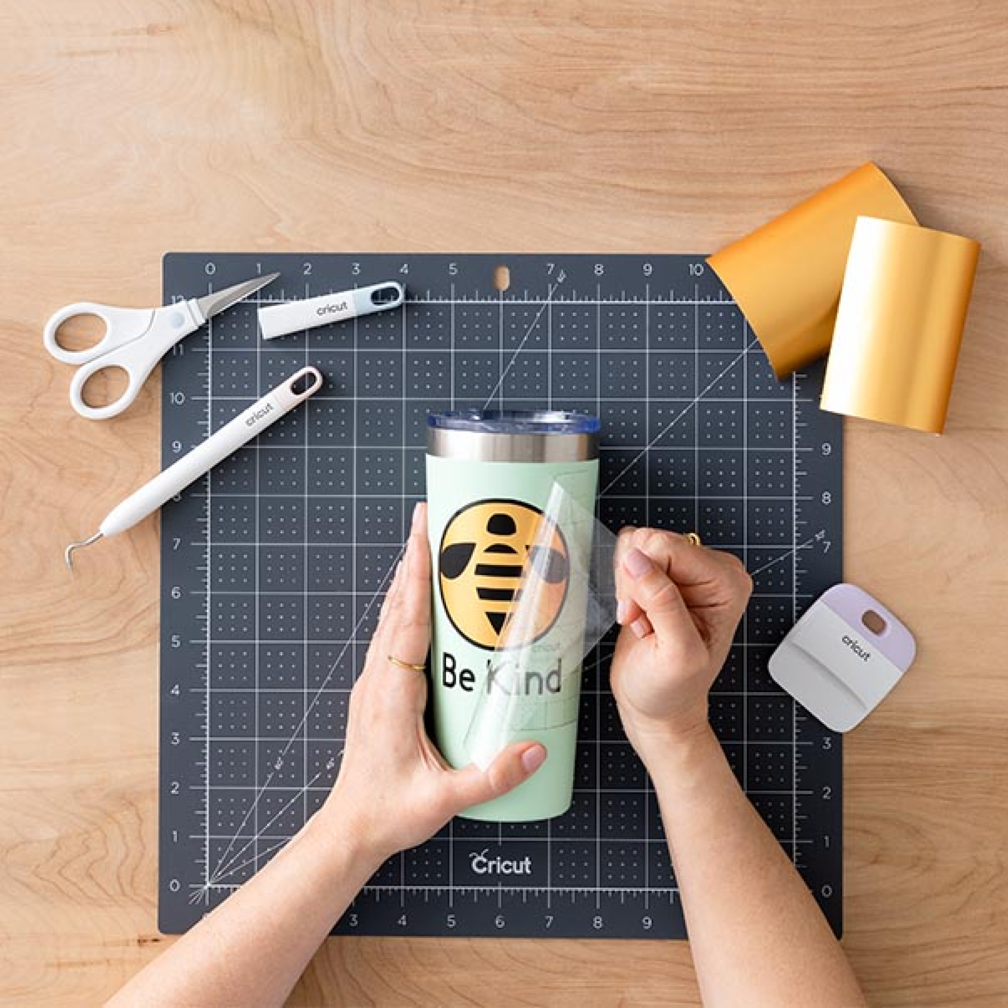 Hände ziehen ein Abziehbild von einem mintgrünen Becher mit einem Bienendesign und den Worten Be Kind ab, umgeben von Bastelwerkzeugen.