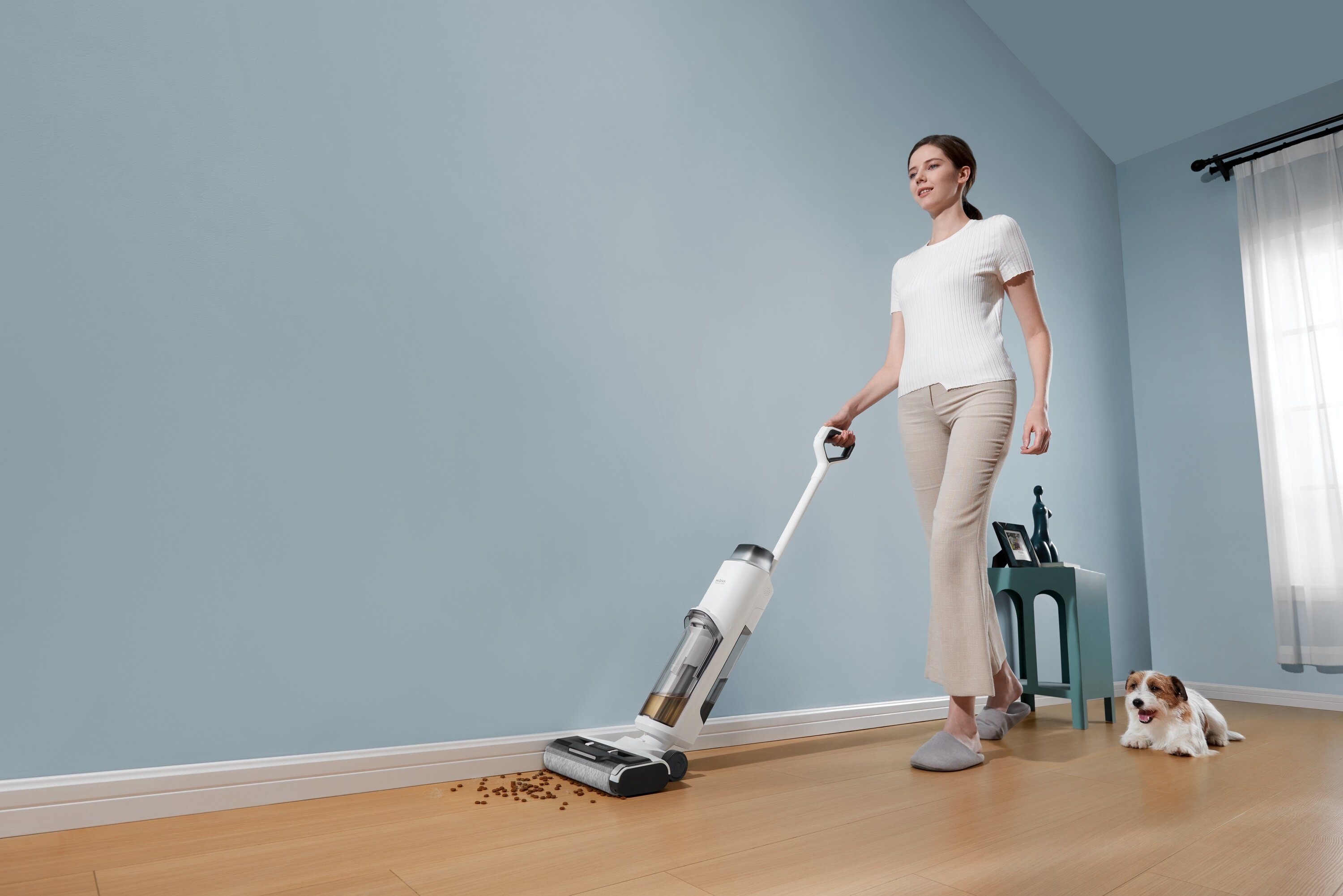 Kobieta odkurza drewnianą podłogę. W pobliżu leży pies. Pokój ma niebieskie ściany i okno z zasłoną.