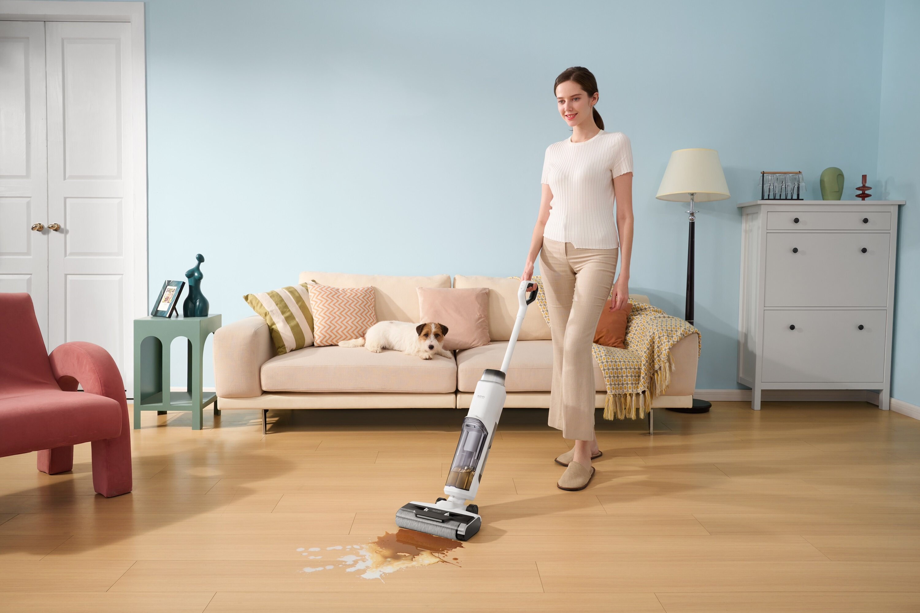 Kobieta odkurza rozlaną ciecz na drewnianej podłodze. Pies odpoczywa na kanapie. W tle lampa i szafka.