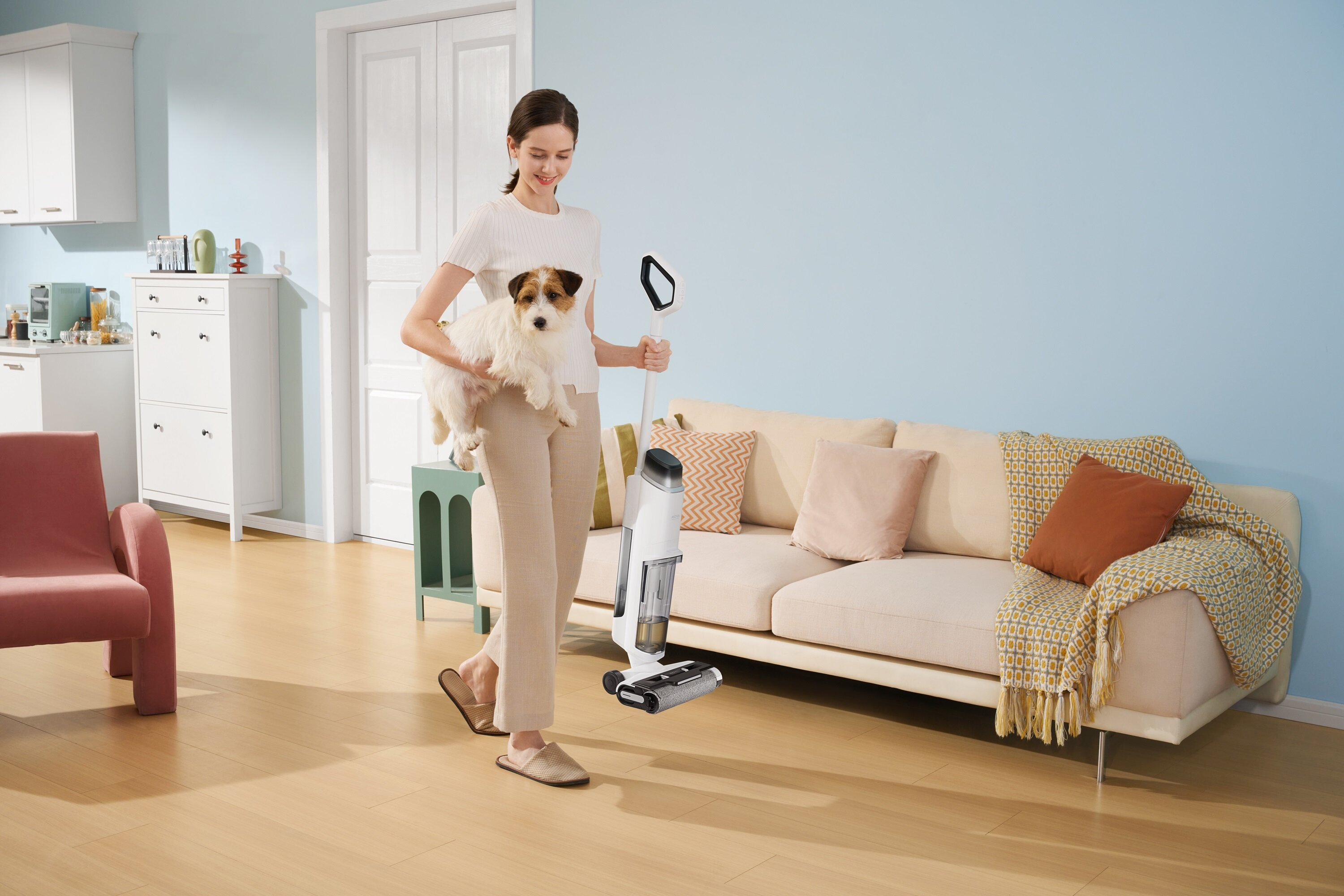 Kobieta trzyma psa i odkurzacz. W tle kanapa i biała szafka. Pokój jest jasnoniebieski.