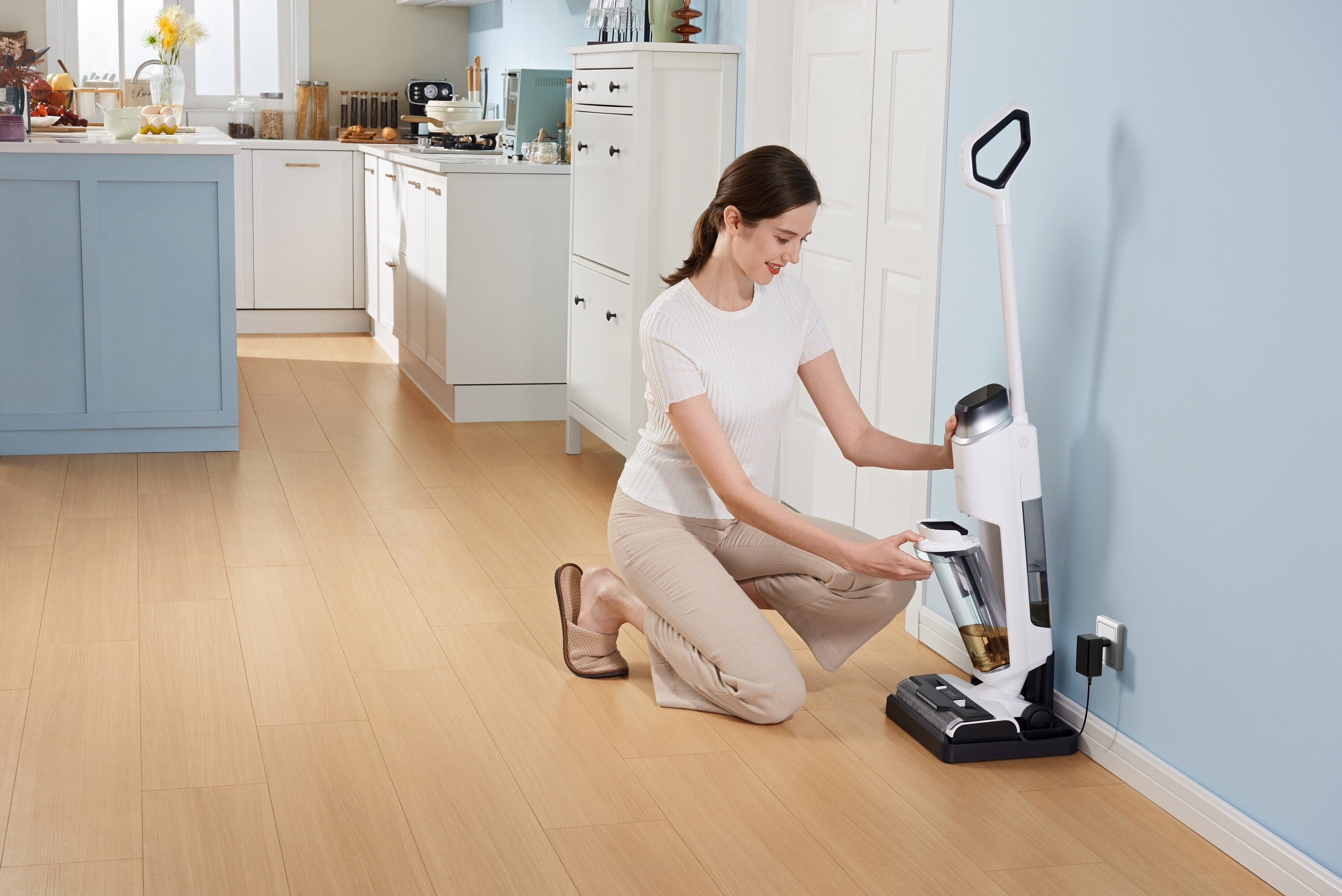 Kobieta klęczy, używając białego i czarnego urządzenia do czyszczenia podłóg. Kuchnia i jasnoniebieska ściana w tle.