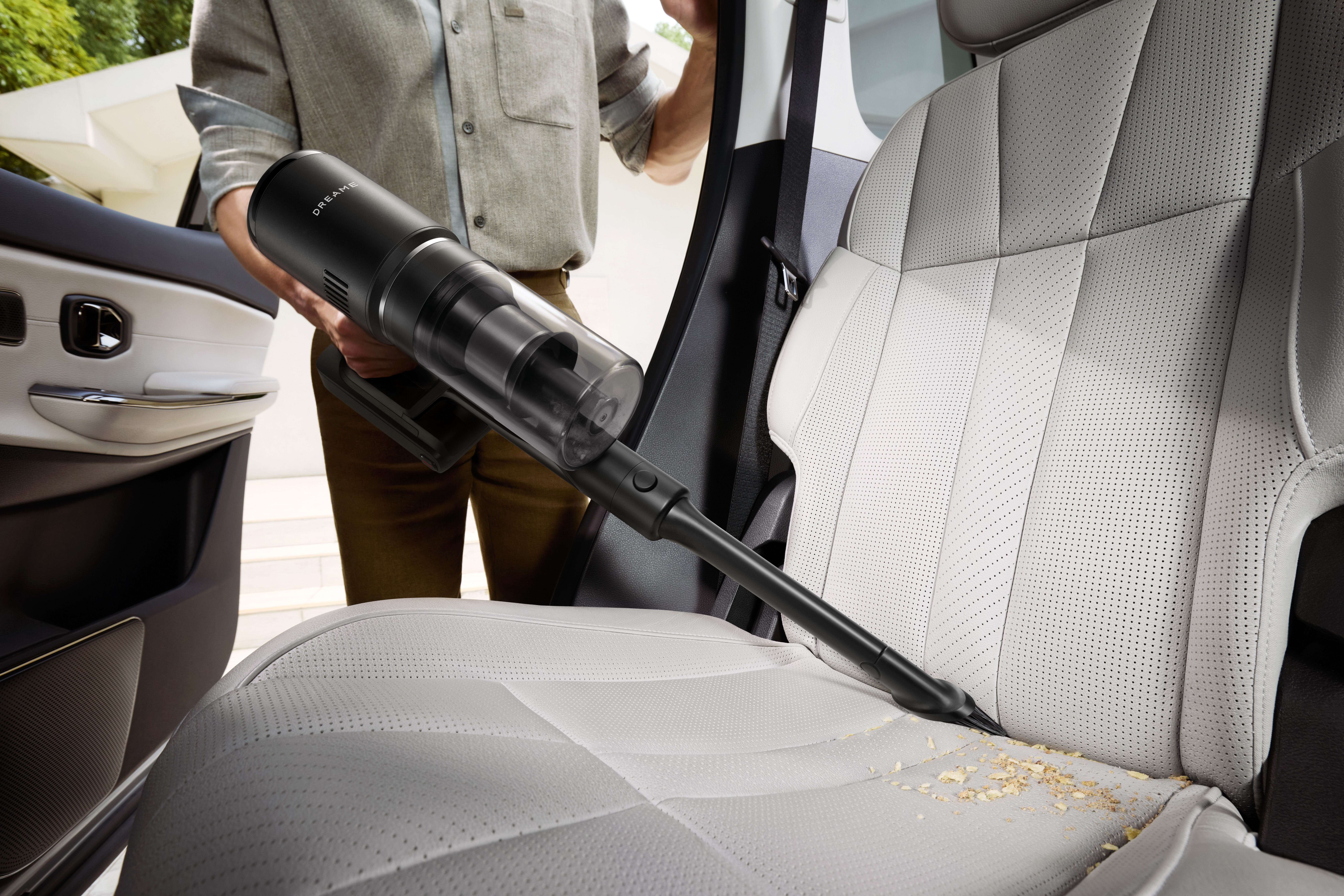 Osoba odkurza fotel samochodowy. Siedzisko jest jasnoszare i ma na sobie zanieczyszczenia. Używana jest czarna odkurzacz.