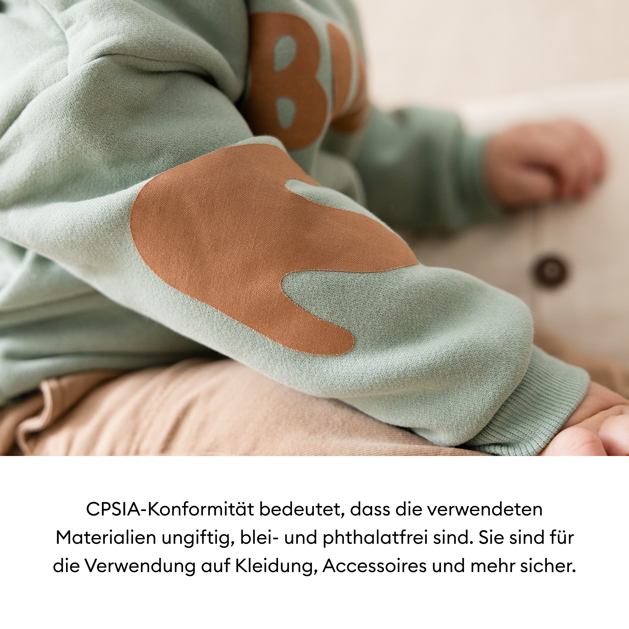 Der Arm eines Kindes, das einen mintgrünen Sweatshirt mit braunen Ellbogenflicken trägt. Der Hintergrund ist verschwommen, neutrale Töne.