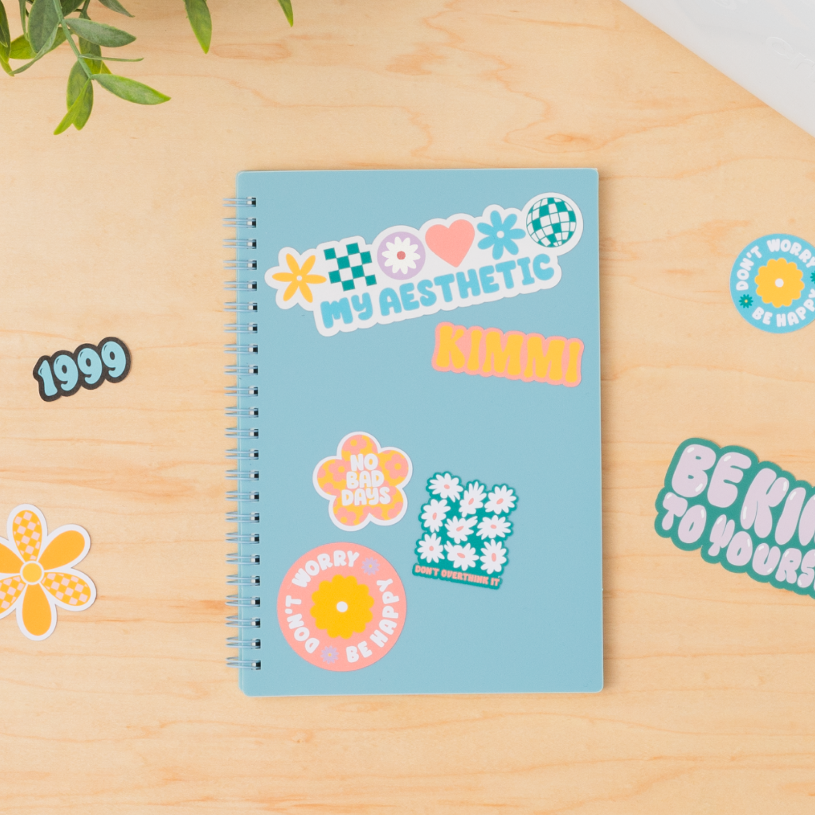 Ein hellblaues Notizbuch mit Aufklebern auf einer Holzoberfläche. Aufkleber sagen 'My Aesthetic', '1999' und Blumendesigns.