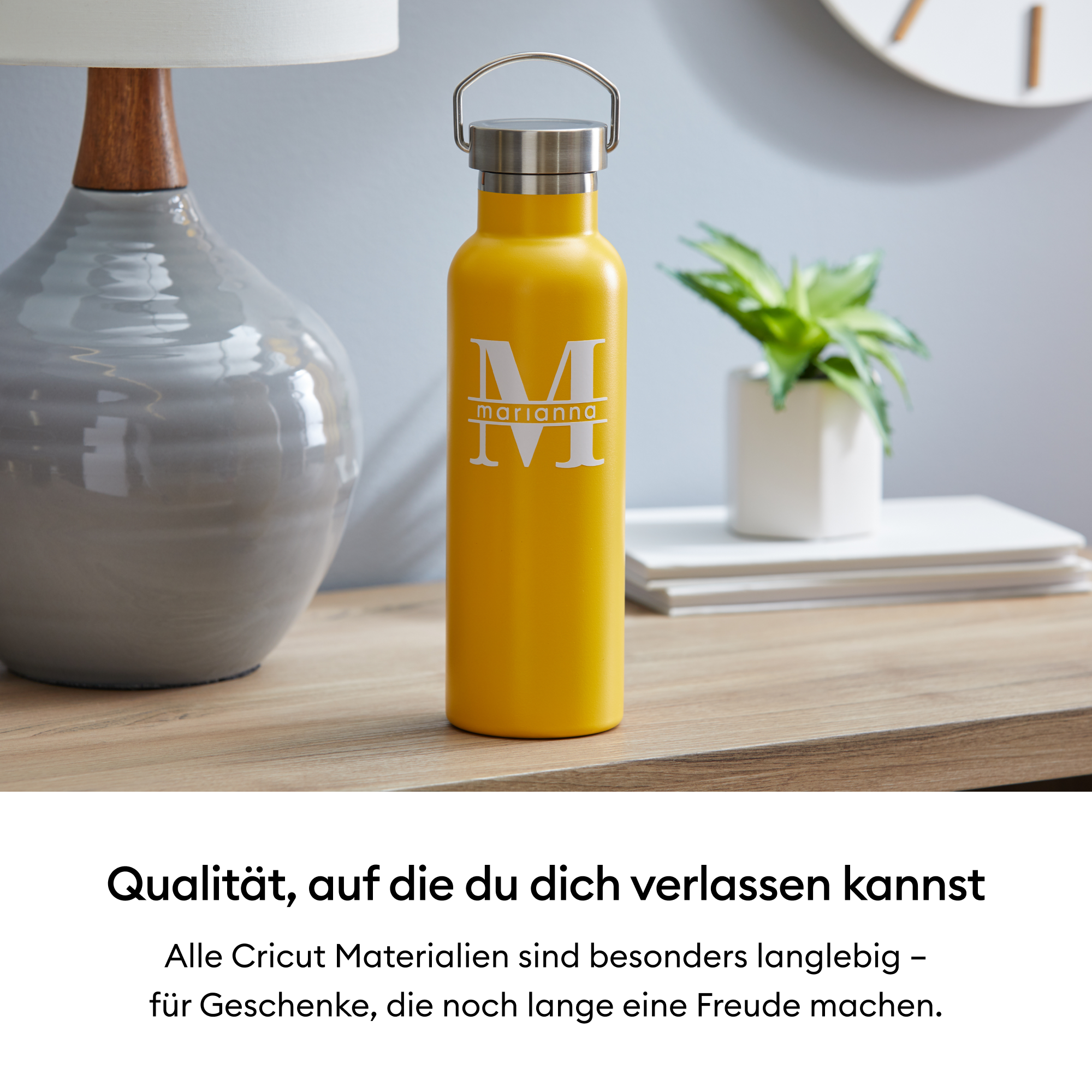Gelbe Wasserflasche mit weißem 'M' und 'marianna' Text, auf einem Holztisch. Eine Lampe und Pflanze im Hintergrund.