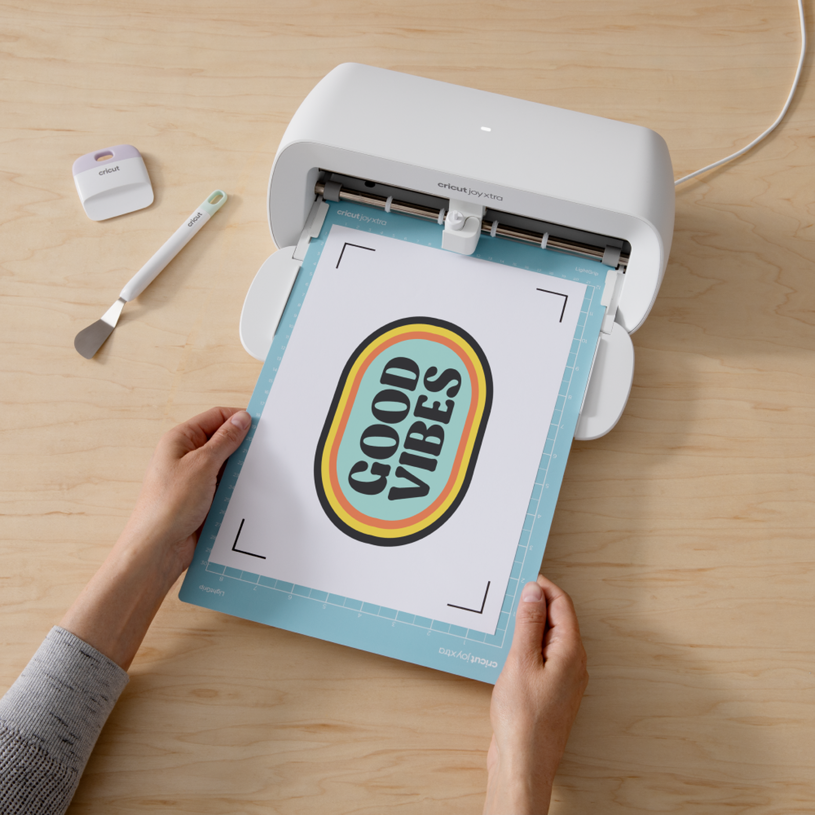 Hände halten eine hellblaue Matte mit einem 'GOOD VIBES'-Aufdruck in einer Cricut Joy Xtra-Maschine auf einer Holzoberfläche.