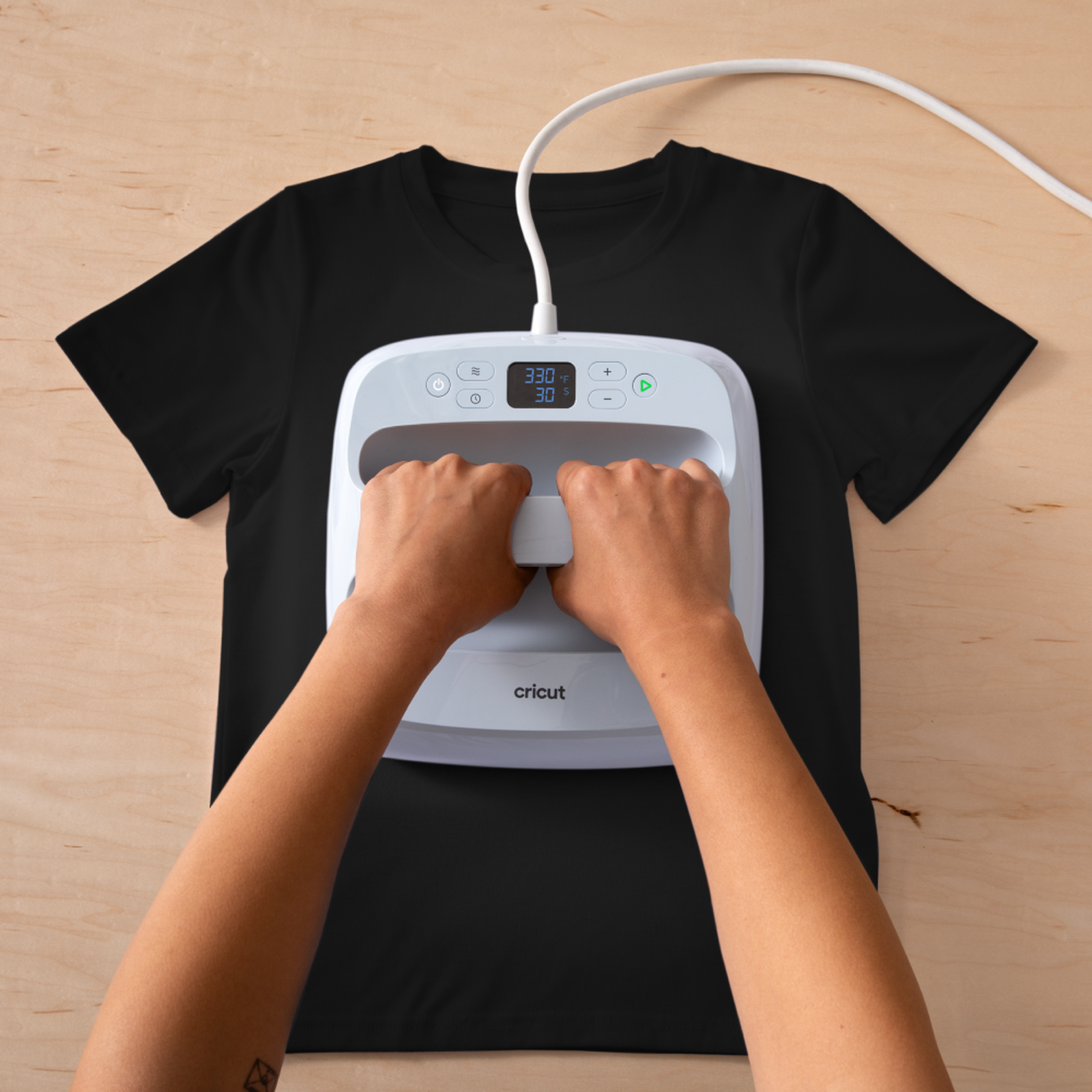 Hände, die eine Cricut-Heißpresse auf einem schwarzen T-Shirt verwenden, das auf einer Holzoberfläche liegt. Die Presse zeigt Temperatur und Zeit an.