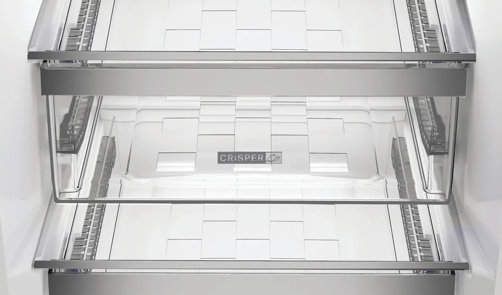 Wnętrze lodówki, prezentujące przezroczyste szuflady na warzywa z metalowymi akcentami. Widoczne logo szuflady. Wnętrze jest czyste, białe.