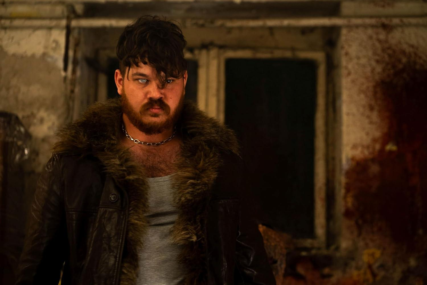 Ein Mann mit dunklen Haaren und Bart trägt eine Lederjacke mit Fell. Er hat einen intensiven Blick und schaut den Betrachter direkt an.