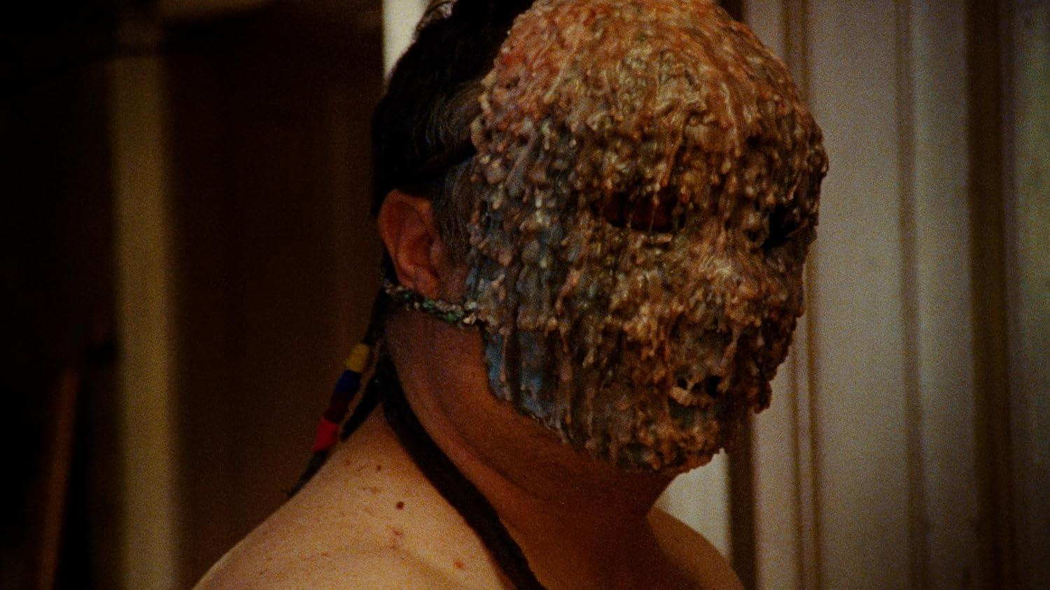 Das Gesicht einer Person ist teilweise von einer Maske aus einer strukturierten, braunen Substanz bedeckt. Dunkle Haare und Haut sind sichtbar.