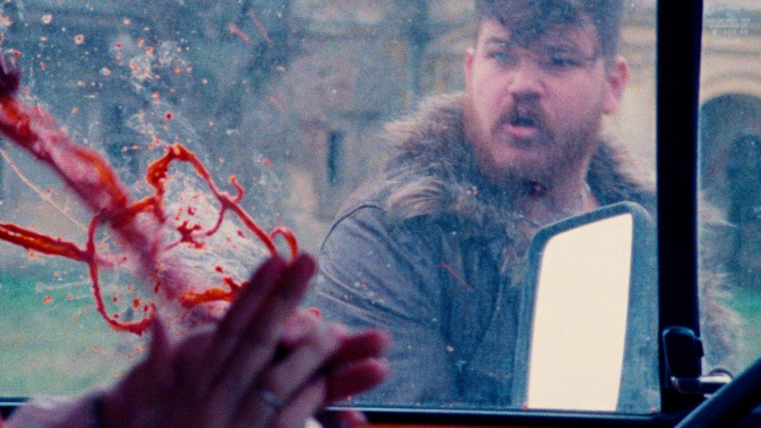 Ein Mann mit Bart blickt aus einem Autofenster, das mit roter Flüssigkeit bespritzt ist. Eine Hand greift zum Glas.