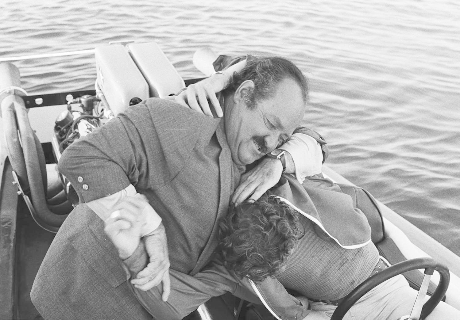Zwei Männer in einem Boot. Einer umarmt den anderen. Beide tragen Anzüge. Der Hintergrund ist das Wasser.