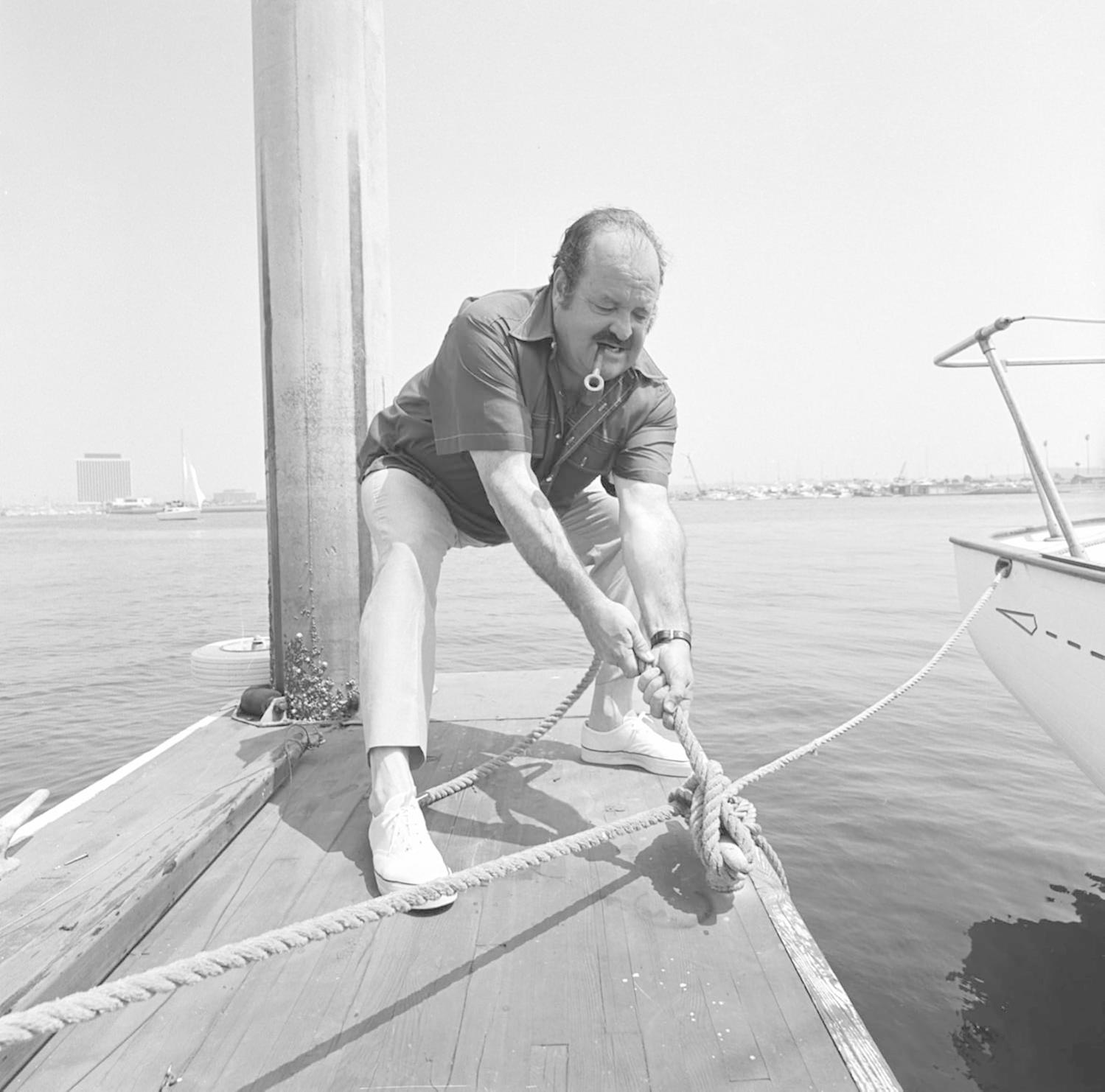 Ein Mann mit Pfeife zieht ein Seil auf einem Holzsteg. Ein Boot ist in der Nähe angebunden. Im Hintergrund Wasser, Boote und Gebäude.