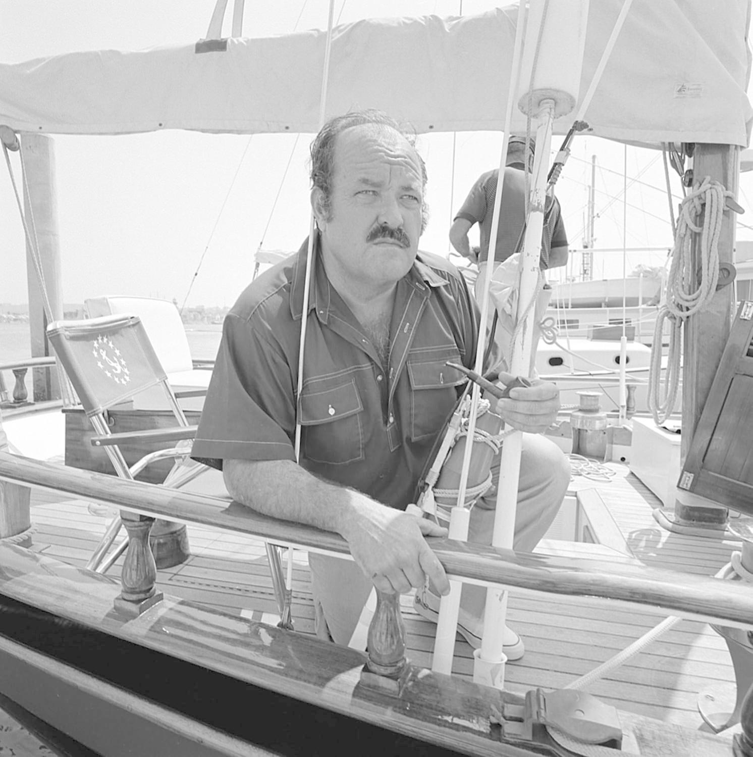 Mann mit Schnurrbart auf einem Boot, Pfeife haltend. Eine andere Person im Hintergrund in der Nähe des Masts. Segelbootdeck im Sonnenlicht.
