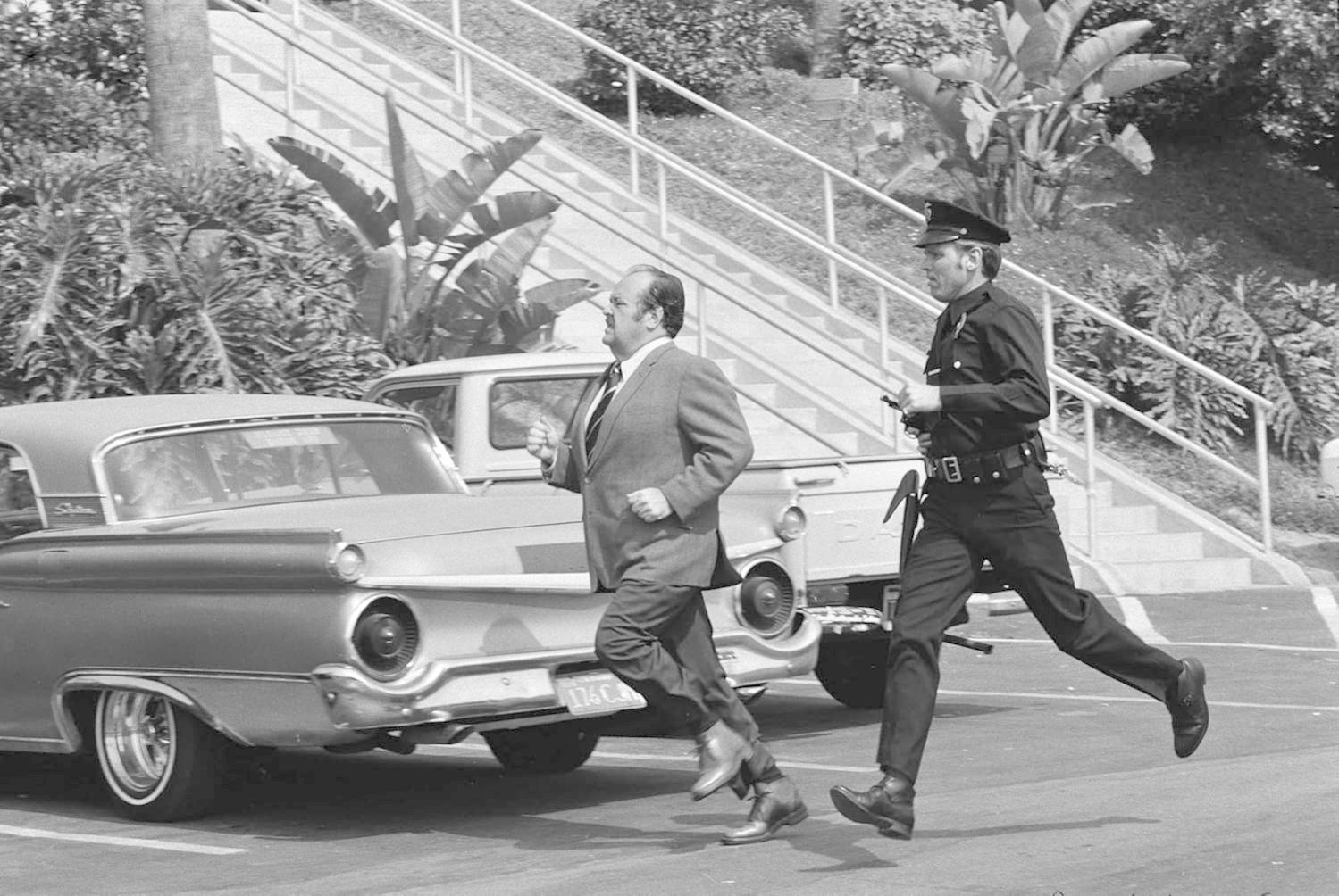 Ein Mann im Anzug rennt vor einem Polizisten in Uniform davon. Autos und Treppen sind im Hintergrund.