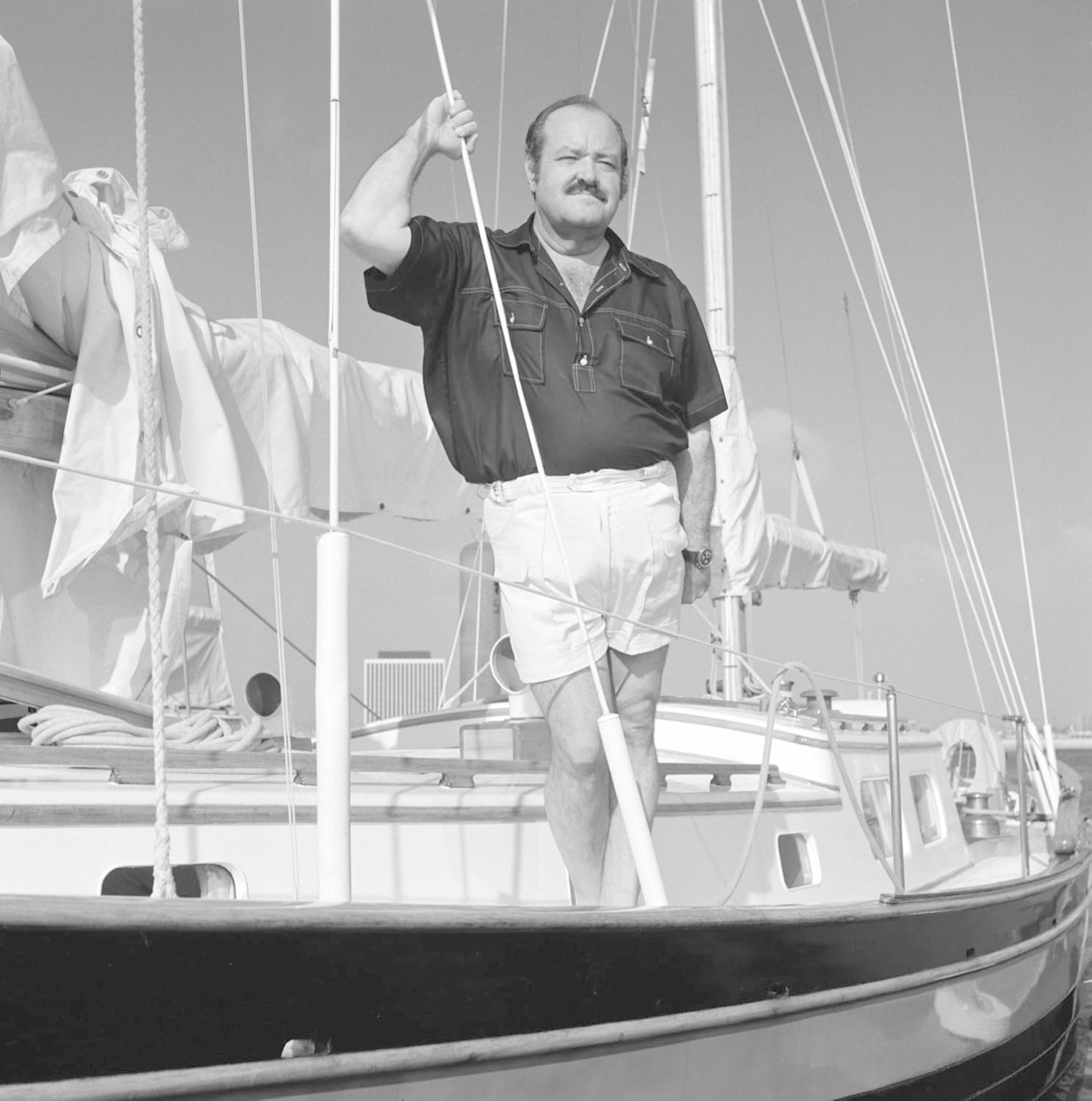 Ein Mann auf einem Segelboot. Er hält ein Seil. Er trägt Shorts und ein Hemd. Der Himmel ist klar.