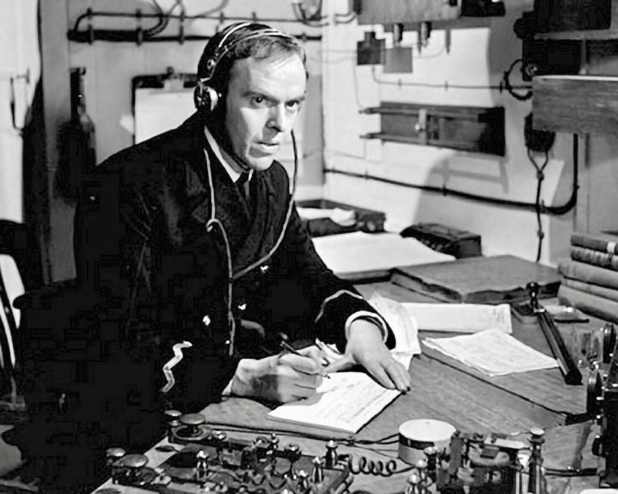 Mann in Marineuniform mit Headset schreibt an einem Schreibtisch mit Funkanlagen, Papieren und Büchern. Szene wirkt vintage.