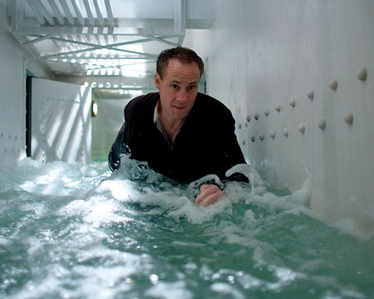 Ein Mann in einem schwarzen Mantel schwimmt durch Wasser in einem weißwandigen Korridor. Das Wasser ist unruhig mit Schaum. Der Mann blickt direkt in die Kamera.