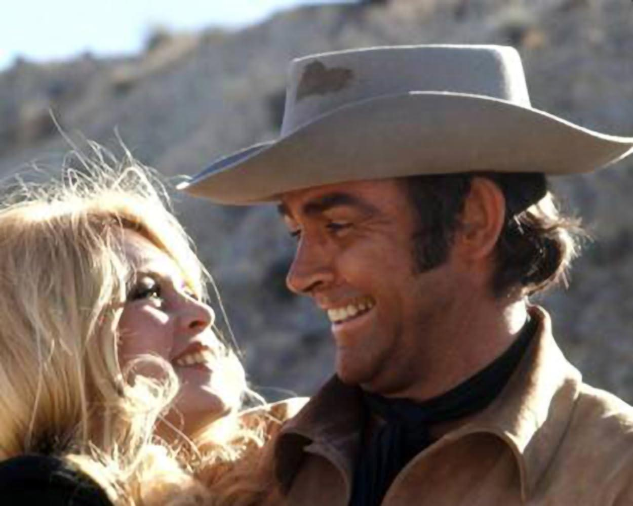 Ein lächelnder Mann mit Cowboyhut blickt eine blonde Frau an. Sie lächeln in einer Umgebung im Freien. Der Hintergrund zeigt eine Wüste.