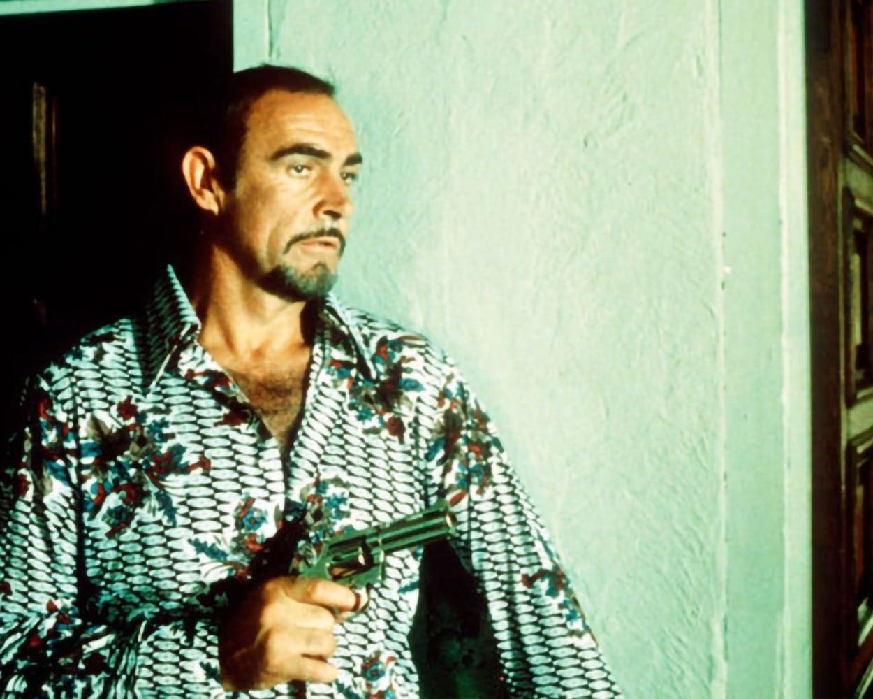 Sean Connery mit einer Waffe, blickt zur Seite. Er trägt ein gemustertes Hemd und steht vor einer hellgrünen Wand.