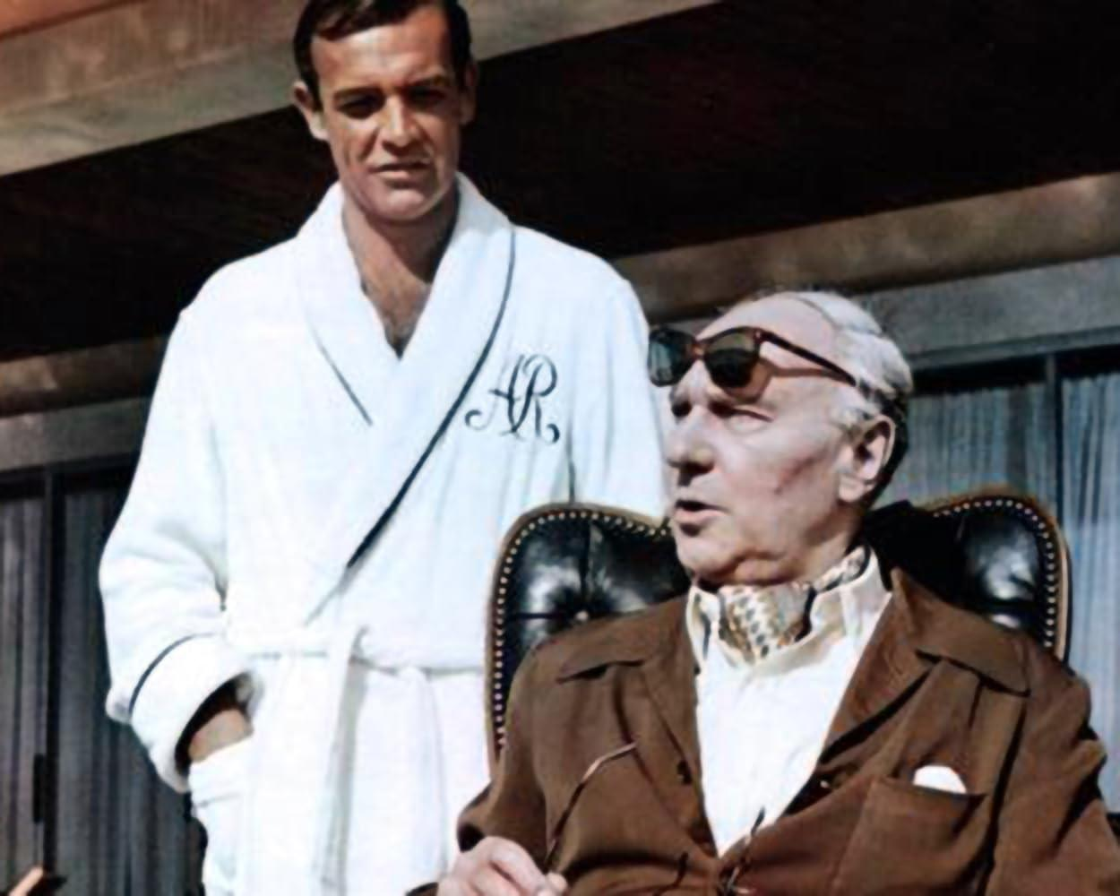 Ein Mann in einem weißen Bademantel steht hinter einem Mann mit Sonnenbrille, der sitzt. Der sitzende Mann trägt eine braune Jacke und einen gemusterten Schal.