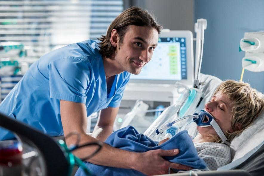 Ein Arzt in blauer OP-Kleidung kümmert sich um einen Patienten in einem Krankenhausbett, der eine Sauerstoffmaske trägt. Im Hintergrund befindet sich ein Monitor.