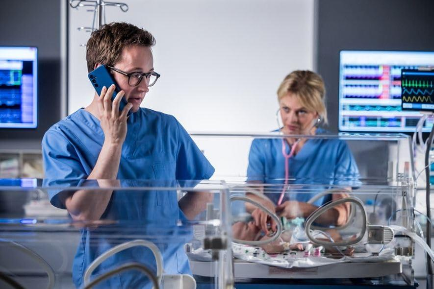 Ein Arzt telefoniert, eine Krankenschwester mit Stethoskop betreut ein Baby im Inkubator. Medizinische Monitore im Hintergrund.