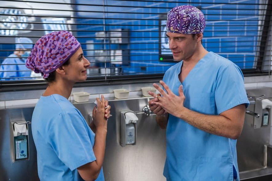 Zwei medizinische Fachkräfte in OP-Kleidung und gemusterten Mützen unterhalten sich. Sie stehen in der Nähe eines Edelstahlbeckens, wahrscheinlich in einem Krankenhaus.