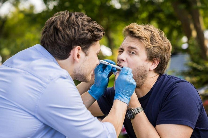 Ein Mann in blauen Handschuhen untersucht den Hals eines anderen Mannes mit einem medizinischen Instrument. Der Hintergrund umfasst grüne Bäume und ein blaues Hemd.