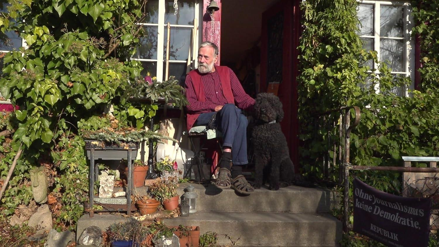 Ein älterer Mann sitzt mit einem schwarzen Hund auf der Veranda. Grüner Efeu bedeckt die Wände. Blumentöpfe sind sichtbar.