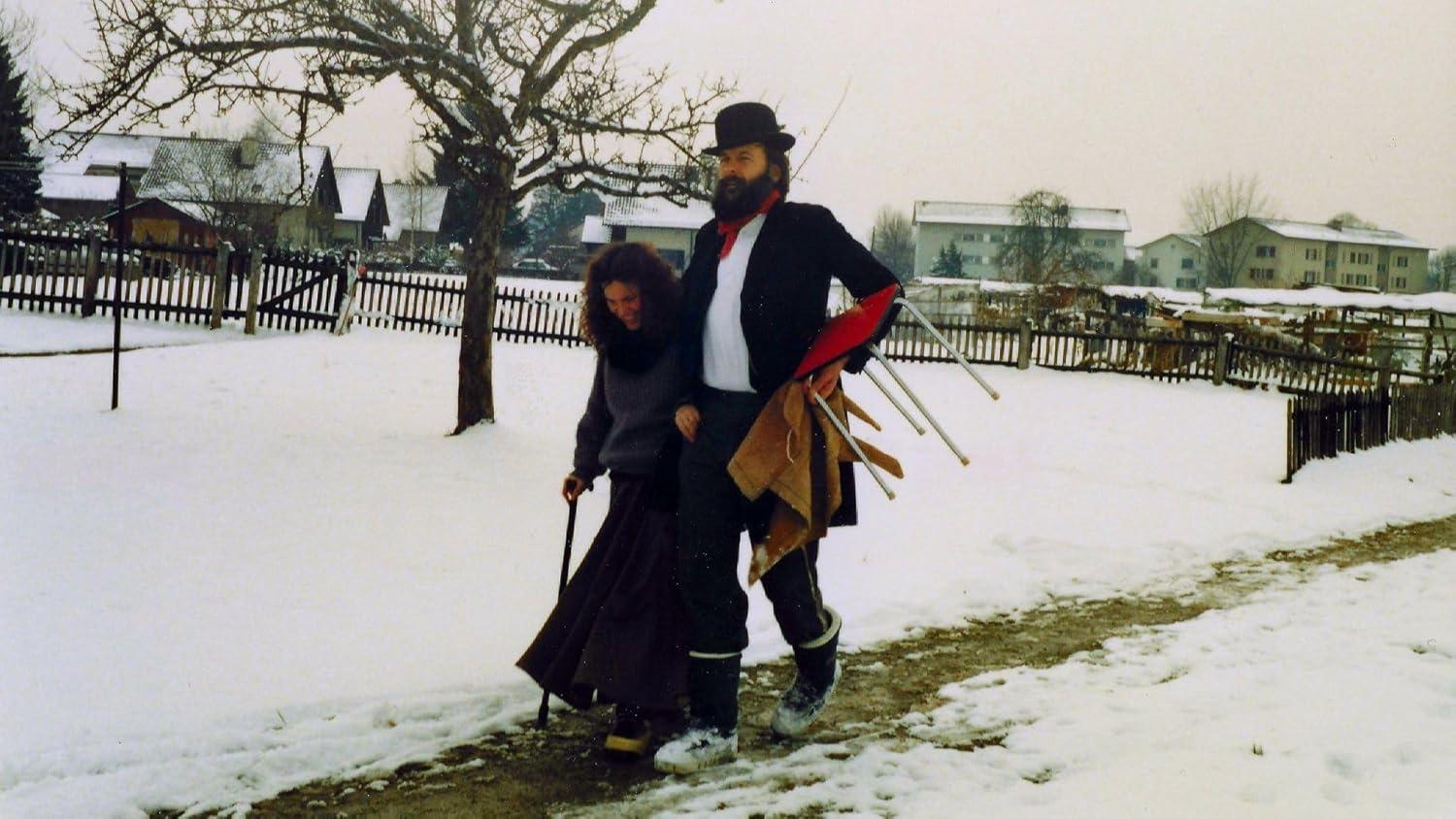 Ein Paar geht im Schnee spazieren. Mann mit Hut hält Stühle und eine Decke. Häuser und ein Zaun im Hintergrund. Winterszene.