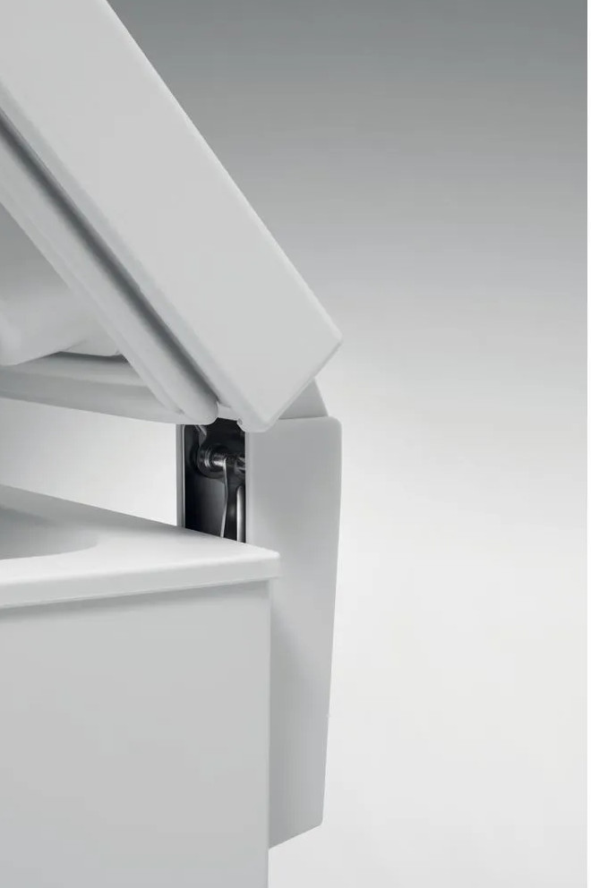 Zbliżenie białej, kanciastej konstrukcji z klapą na zawiasach. Widoczny jest metalowy zawias. Tło to gradient bieli.