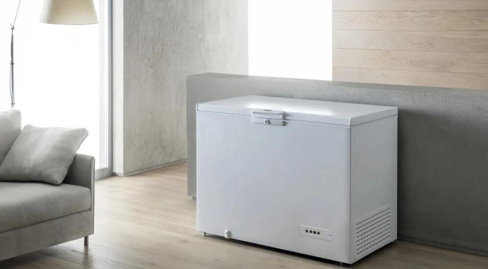 Biała zamrażarka skrzyniowa w pokoju z sofą, lampą i drewnianą podłogą. Szare ściany i okno w tle.